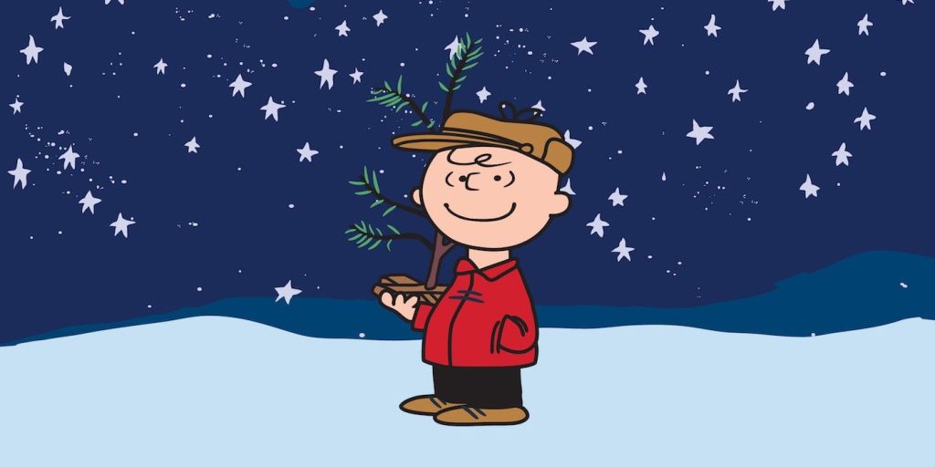 A still from the animated TV Show "A Charlie Brown Christmas" from 1965. In this image, the titular character, a small boy named Charlie Brown is outside, wearing a red jacket, black pants, brown shoes and a brown "trappers" hat. He is holding a small, scraggly pine branch as a Christmas tree. He is standing on snow, smiling, staring at a star filled night sky.